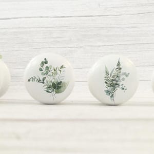 K&ouml;nnte beinhalten: Vier wei&szlig;e Keramik-Schrankkn&ouml;pfe mit botanischen Designs. Jeder Knopf zeigt eine andere Anordnung von gr&uuml;nen Bl&auml;ttern und Stielen. Die Kn&ouml;pfe sind in einer Reihe vor einem wei&szlig;en Holzgrund angeordnet.