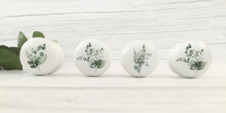 K&ouml;nnte beinhalten: Vier wei&szlig;e Keramik-Schrankkn&ouml;pfe mit botanischen Designs. Jeder Knopf zeigt eine andere Anordnung von gr&uuml;nen Bl&auml;ttern und Stielen. Die Kn&ouml;pfe sind in einer Reihe vor einem wei&szlig;en Holzgrund angeordnet.
