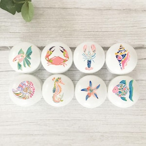 May include: Eight white ceramic cabinet knobs with colourful sea life designs. The knobs feature illustrations of a goldfish, crab, lobster, seashell, nautilus, seahorse, starfish, and another fish. The knobs are arranged on a light wood surface.