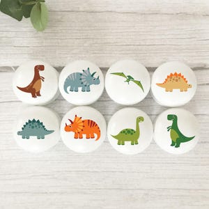 May include: Eight white cabinet knobs, each decorated with a different dinosaur illustration. The dinosaurs are depicted in various colours, including brown, blue, orange, green, and yellow. The knobs are arranged in two rows of four.