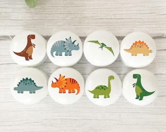 May include: Eight white cabinet knobs with colorful dinosaur illustrations. Each knob features a different dinosaur design in shades of brown, blue, orange, green, and yellow. The knobs are arranged in two rows of four.