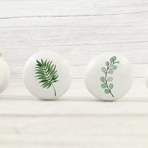 K&ouml;nnte beinhalten: Vier wei&szlig;e Keramikkn&ouml;pfe mit gr&uuml;nen Blattdesigns. Die Kn&ouml;pfe sind rund und haben eine glatte, gl&auml;nzende Oberfl&auml;che.