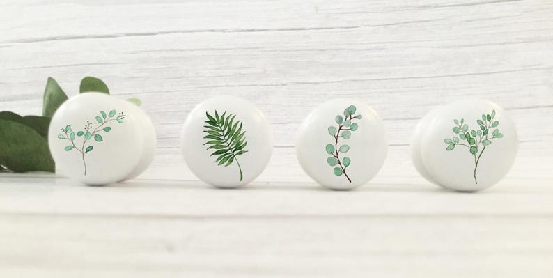K&ouml;nnte beinhalten: Vier wei&szlig;e Keramikkn&ouml;pfe mit gr&uuml;nen Blattdesigns. Die Kn&ouml;pfe sind rund und haben eine glatte, gl&auml;nzende Oberfl&auml;che.