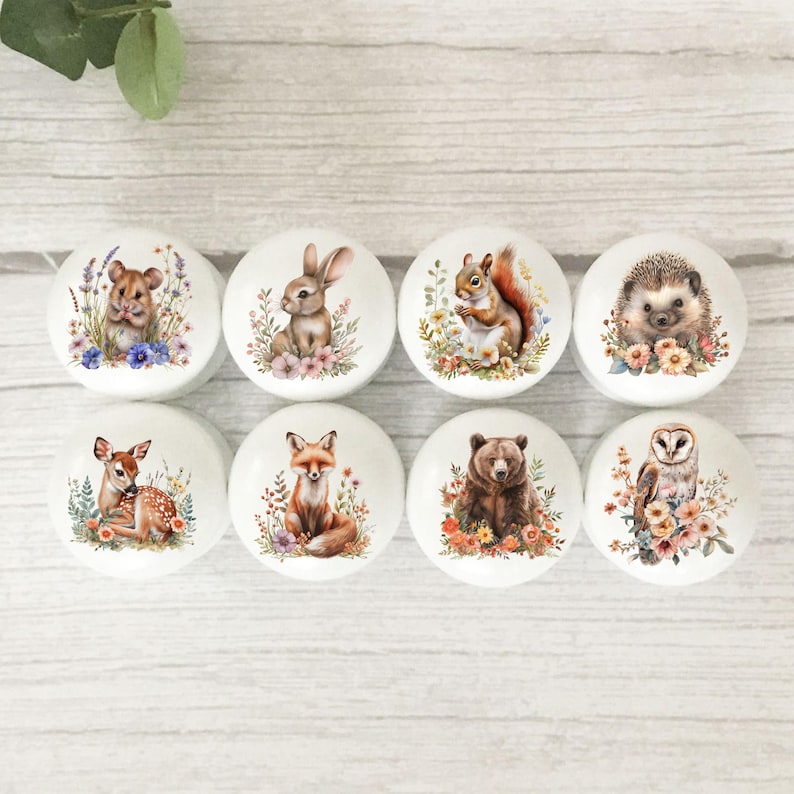 May include: Eight white ceramic cabinet knobs with woodland animal illustrations. Each knob depicts a different animal: a hamster, rabbit, squirrel, hedgehog, deer, fox, bear, and owl, surrounded by floral details. The knobs are displayed on a light wood surface.