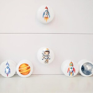 Op de afbeelding: Set van 7 ronde witte ladeknopjes met ruimtethema illustraties. De knoppen tonen een astronaut, planeten en raketten.