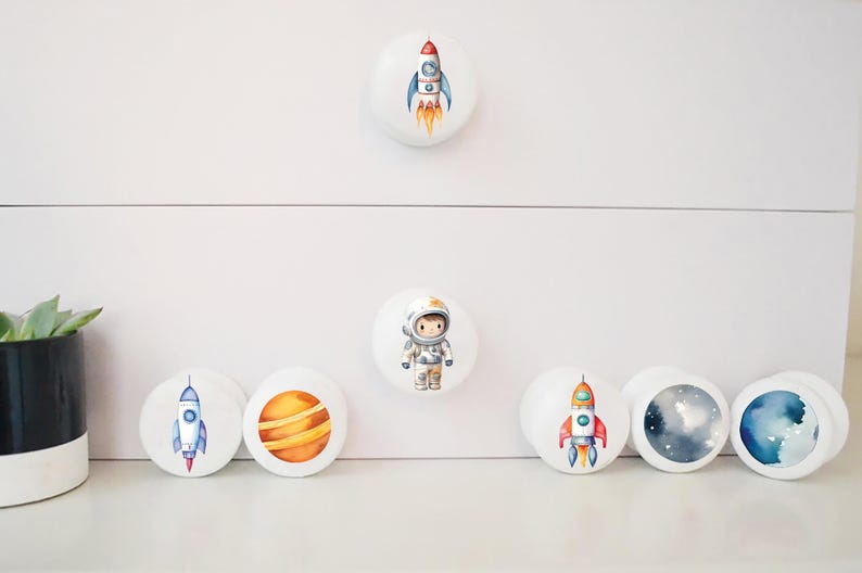 Op de afbeelding: Set van 7 ronde witte ladeknopjes met ruimtethema illustraties. De knoppen tonen een astronaut, planeten en raketten.