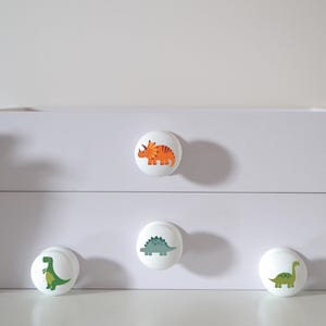 Four white solid Beech furniture knobs with colourful dinosaur designs. The dinosaurs include an orange Triceratops, a blue/grey Stegosaurus, a green T Rex and a green Brachiosaurus. The drawer handles have a smooth glossy finish and are shown on a white chest of drawers.