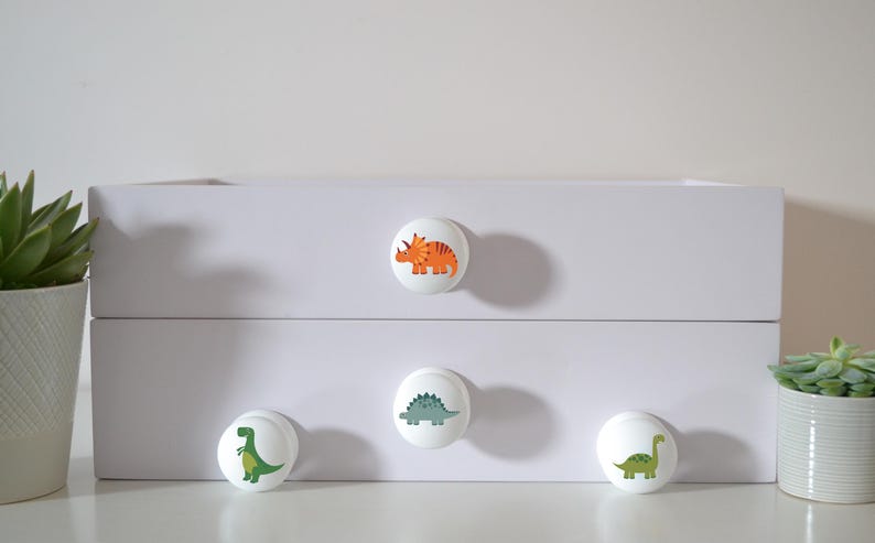 Four white solid Beech furniture knobs with colourful dinosaur designs. The dinosaurs include an orange Triceratops, a blue/grey Stegosaurus, a green T Rex and a green Brachiosaurus. The drawer handles have a smooth glossy finish and are shown on a white chest of drawers.