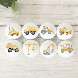 May include: Eight white ceramic cabinet knobs with yellow construction vehicle designs. The knobs feature illustrations of a crane truck, dumper truck, front-end loader, cement mixer, excavator, and a tractor.