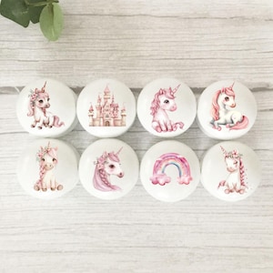 May include: Eight white cabinet knobs with unicorn and castle designs. The knobs feature pink and purple unicorns, a pink castle, and a rainbow. The knobs are round and have a glossy finish.