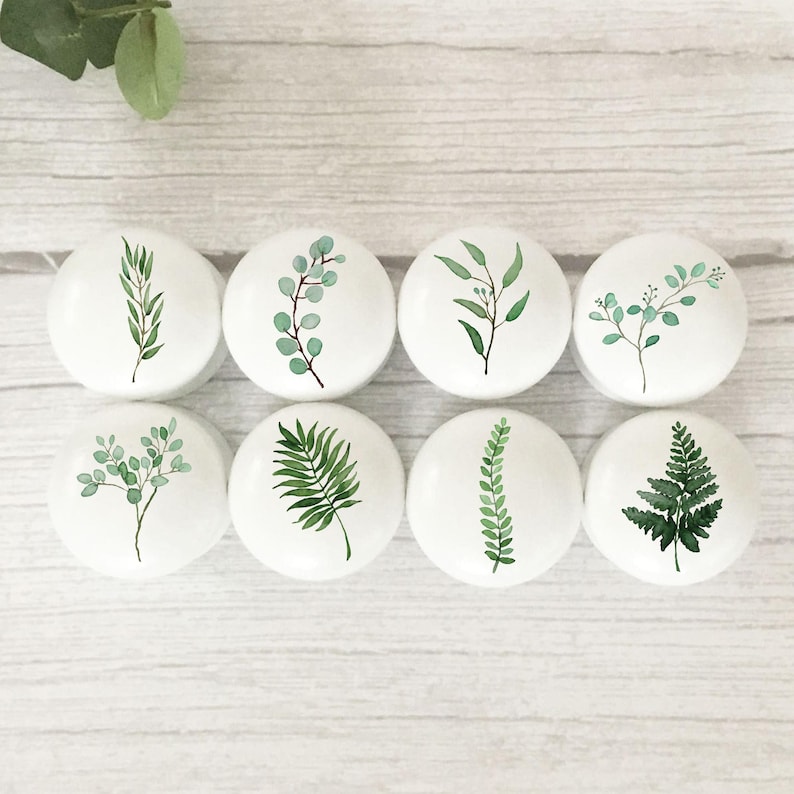 May include: Set of eight white ceramic knobs with green watercolour leaf designs. The knobs feature various leaf shapes, including ferns, eucalyptus, and palm leaves.
