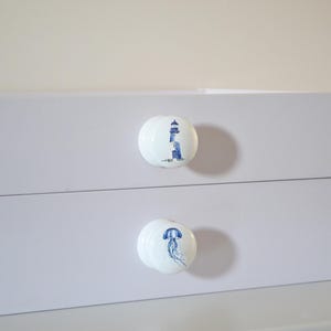 Two white round solid beech furniture knobs each with a different nautical design. The designs include a lighthouse and a jellyfish. The cabinet knobs have a glossy finish and are shown on a chest of drawers with succulent plants for decoration.