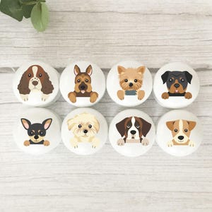May include: Eight white ceramic cabinet knobs with various dog breeds illustrated on them. The knobs depict a Spaniel, German Shepherd, Yorkshire Terrier, Rottweiler, Chihuahua, Maltese, Saint Bernard, and Jack Russell Terrier. Each knob has a detailed dog face.