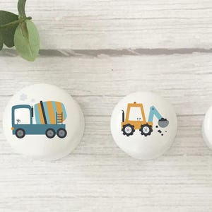 Three white round solid beech furniture knobs each with a different construction vehicle design on. They include a cement mixer, digger and crane. The colour scheme is a teal blue, mustard and brown. The handles have a glossy finish and are shown in 3 sizes shown from the largest to the smallest.