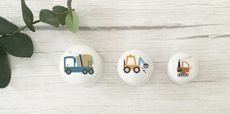 Three white round solid beech furniture knobs each with a different construction vehicle design on. They include a cement mixer, digger and crane. The colour scheme is a teal blue, mustard and brown. The handles have a glossy finish and are shown in 3 sizes shown from the largest to the smallest.