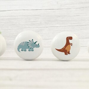 Four white solid Beech cabinet knobs with dinosaur illustrations. The knobs feature a green Brontosaurus, a blue Triceratops, a brown T-Rex, and a blue Stegosaurus. These decorative knobs are perfect for a child's room or nursery.