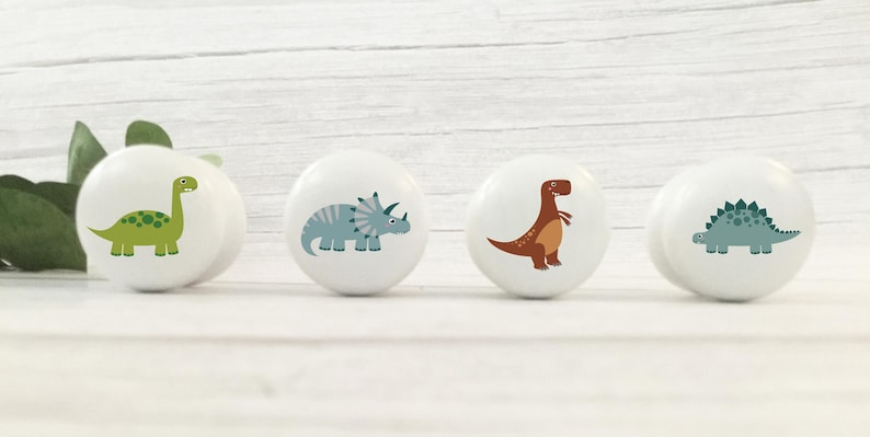 Four white solid Beech cabinet knobs with dinosaur illustrations. The knobs feature a green Brontosaurus, a blue Triceratops, a brown T-Rex, and a blue Stegosaurus. These decorative knobs are perfect for a child's room or nursery.