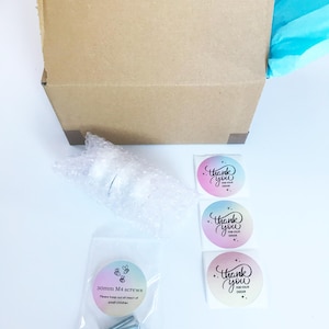 May include: A cardboard box with blue tissue paper filling. The box contains a white knob, three silver screws, and three thank you stickers with a rainbow gradient background. The stickers say "Thank you for your order".