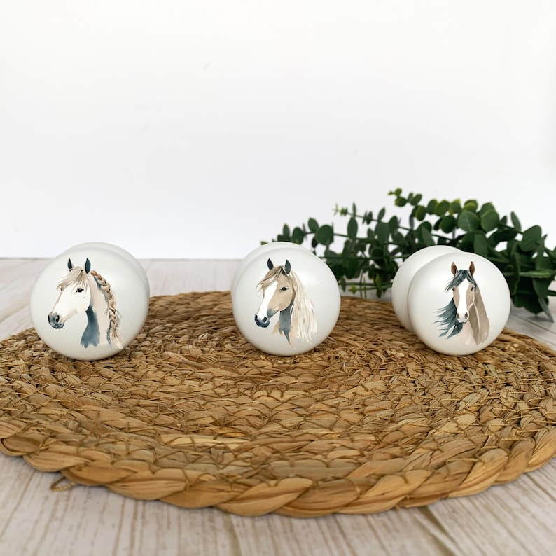 Three white round solid beech furniture knobs with different horse head illustrations. The horses consist of a white/tan, a beige/tan and a light brown one. The drawer pulls have a glossy finish and are shown on a pale wood background with a wicker mat and foliage for decoration.