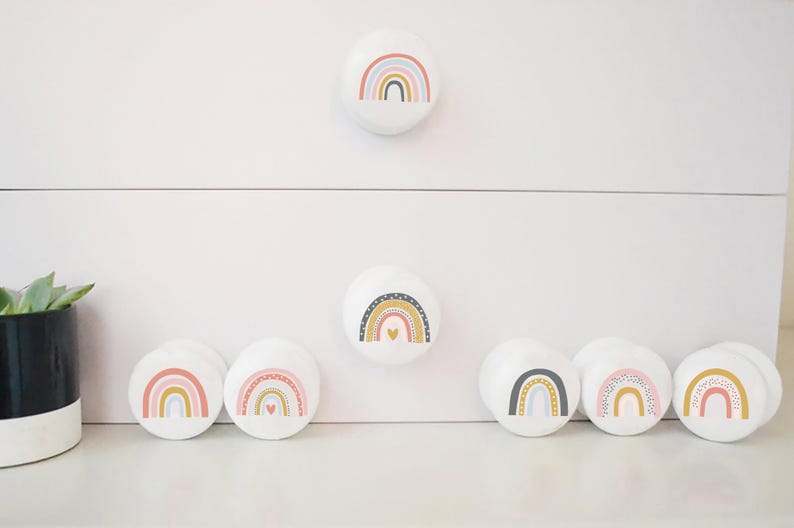 Peut inclure: Sept boutons de tiroir ronds blancs avec des motifs arc-en-ciel. Les arcs-en-ciel sont de diff&eacute;rentes couleurs, notamment le rose, le bleu, le jaune et l'or. Les boutons sont dispos&eacute;s en rang&eacute;e sur une surface blanche.