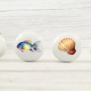 May include: Four white ceramic knobs with different ocean-themed designs. The knobs feature a seal, a fish, a seashell, and a whale.
