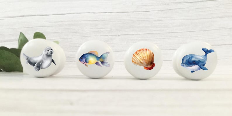 May include: Four white ceramic knobs with different ocean-themed designs. The knobs feature a seal, a fish, a seashell, and a whale.