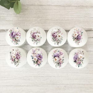 May include: A set of eight round white cabinet knobs, each featuring a printed floral arrangement of purple, pink, and white flowers with green foliage. The knobs are arranged in two rows of four, displaying a consistent botanical design.