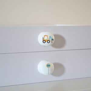 A white chest of drawers with two drawers, each with a round knob. The top knob has an excavator, the bottom a blue stop sign. A small potted plant is to the right for decoration.