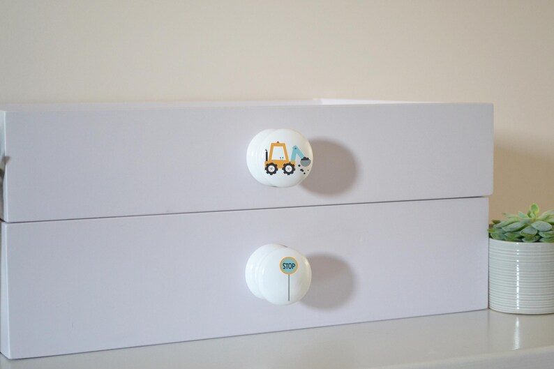 A white chest of drawers with two drawers, each with a round knob. The top knob has an excavator, the bottom a blue stop sign. A small potted plant is to the right for decoration.