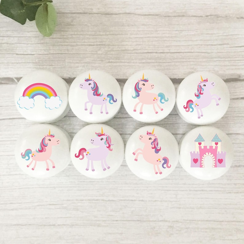 May include: Eight white cabinet knobs featuring unicorn and rainbow designs. The unicorns have pink, purple, and blue manes and tails. One knob shows a rainbow, another a pink castle with hearts.
