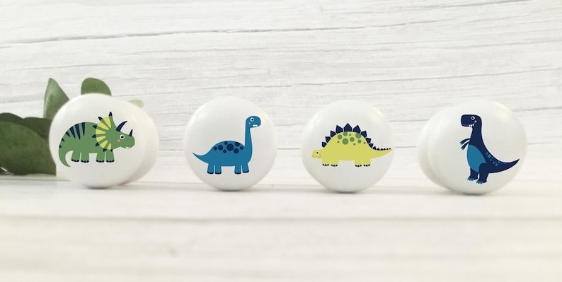 以下が含まれることがあります： 異なる恐竜のデザインが施されたセラミック製のノブが4つ。ノブは白で、青、緑、黄色の恐竜が描かれています。
