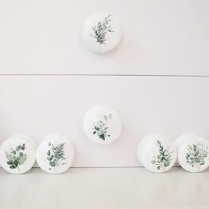 K&ouml;nnte beinhalten: Wei&szlig;e Keramik-Schrankkn&ouml;pfe mit botanischen Designs mit gr&uuml;nen Bl&auml;ttern und Stielen. Die Kn&ouml;pfe sind rund und unterschiedlich gro&szlig;, einige sind auf einer wei&szlig;en Oberfl&auml;che platziert. Eine Topfpflanze ist links zu sehen.