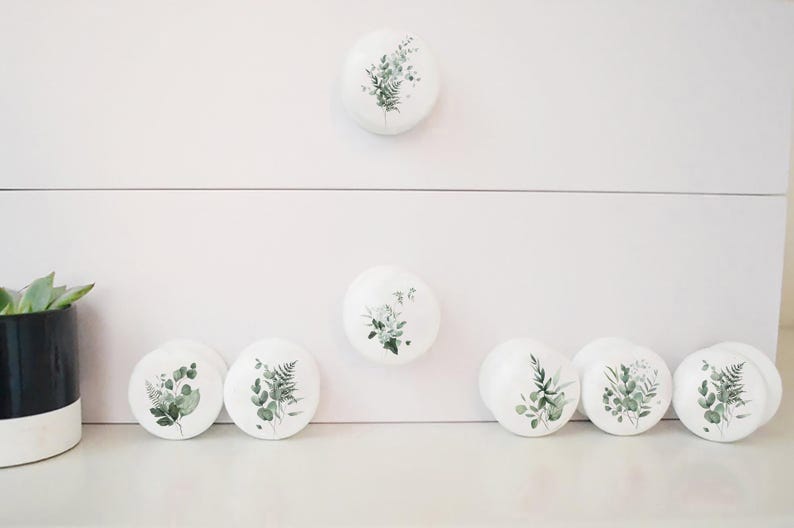 K&ouml;nnte beinhalten: Wei&szlig;e Keramik-Schrankkn&ouml;pfe mit botanischen Designs mit gr&uuml;nen Bl&auml;ttern und Stielen. Die Kn&ouml;pfe sind rund und unterschiedlich gro&szlig;, einige sind auf einer wei&szlig;en Oberfl&auml;che platziert. Eine Topfpflanze ist links zu sehen.