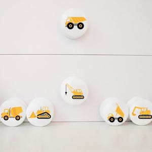 Op de afbeelding: Set van zeven witte ronde trekknopjes voor lades met gele ontwerpen van bouwvoertuigen. De ontwerpen omvatten een kiepwagen, een kraan, een bulldozer, een betonmixer en een graafmachine.