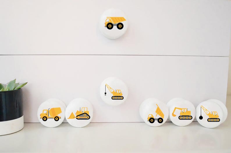 Op de afbeelding: Set van zeven witte ronde trekknopjes voor lades met gele ontwerpen van bouwvoertuigen. De ontwerpen omvatten een kiepwagen, een kraan, een bulldozer, een betonmixer en een graafmachine.