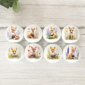 May include: A set of eight round white cabinet knobs, each featuring a unique watercolour illustration of a fox sitting among various colourful wildflowers, including lavender, poppies, and yellow blossoms, arranged in two rows of four.