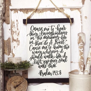 Peut inclure: Une pancarte en toile blanche avec un texte calligraphié noir qui dit "Cause me to hear thy lovingkindness in the morning: for in thee do I trust: Cause me to know the way wherein I should walk: for I lift up my soul unto thee. Psalm 143:8". La pancarte est suspendue à une corde et a un cadre en bois noir.
