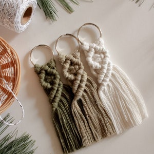 Peut inclure: Trois porte-clés en macramé avec une frange en pompon. Les porte-clés sont dans des tons de vert, beige et blanc.