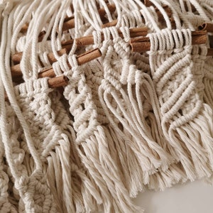 May include: Close-up of a macrame wall hanging in a natural, off-white hue. The macrame features detailed knotting patterns and is accented with cinnamon sticks. The bottom edge has a fringe.