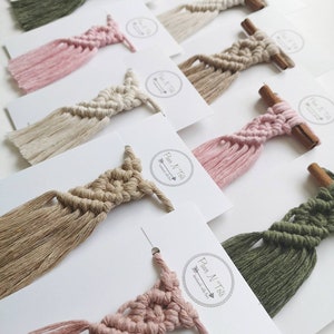 Puede incluir: Una colecci&oacute;n de peque&ntilde;as tapices de macram&eacute; de pared con borlas en varios colores, incluyendo rosa, verde, beige y marr&oacute;n. Cada tapiz est&aacute; unido a una peque&ntilde;a rama de canela.