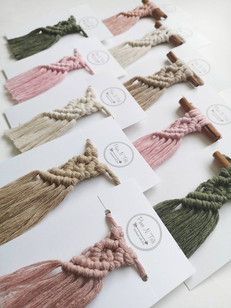 Puede incluir: Una colecci&oacute;n de peque&ntilde;as tapices de macram&eacute; de pared con borlas en varios colores, incluyendo rosa, verde, beige y marr&oacute;n. Cada tapiz est&aacute; unido a una peque&ntilde;a rama de canela.