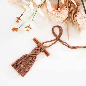 Puede incluir: Un borla de macram&eacute; marr&oacute;n colgando de dos palitos de canela. La borla est&aacute; hecha con un cord&oacute;n grueso marr&oacute;n y tiene una franja en la parte inferior. Los palitos de canela est&aacute;n atados juntos con el cord&oacute;n y cuelgan de un fondo blanco.