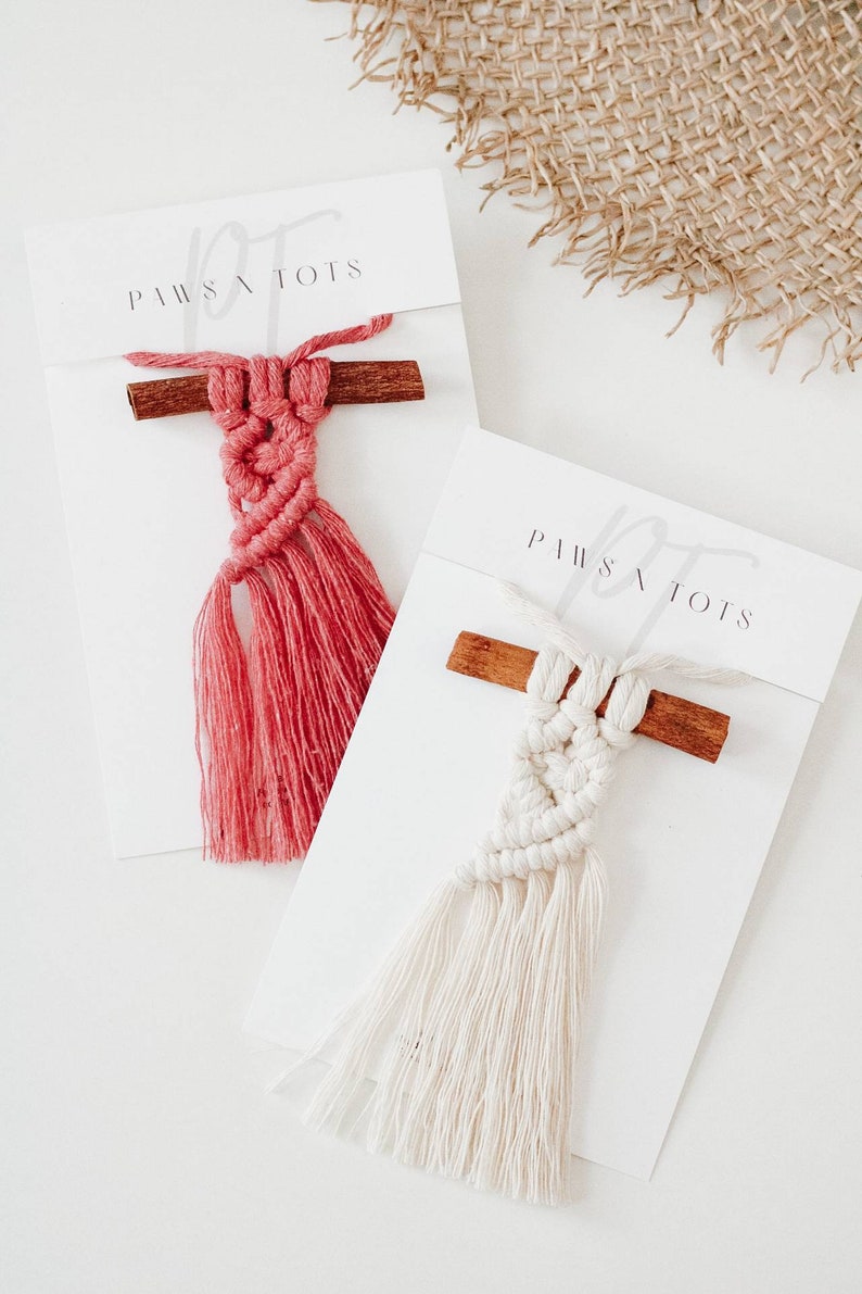 Puede incluir: Dos tapices de macram&eacute; con listones de madera y borlas. Los tapices est&aacute;n sobre tarjetas blancas con el texto "Paws & Tots" impreso en ellas. Un tapiz es rojo y el otro es blanco.