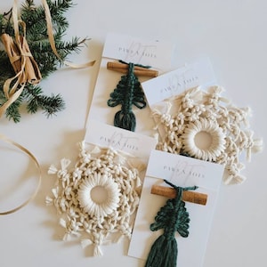 May include: Three handmade macrame ornaments, a snowflake and two Christmas trees, with cinnamon sticks attached. The ornaments are on white cards with the text "PAWS & TOYS" printed on them.