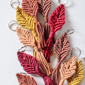 May include: A collection of colourful macrame leaf keychains with tassels. The keychains are made with a variety of colours, including red, orange, yellow, and brown.