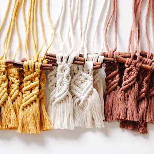 Peut inclure: Une collection de sept tentures murales en macramé faites à la main avec des glands de différentes couleurs, notamment noir, jaune, blanc et marron. Chaque tenture est fixée à une baguette en bois.