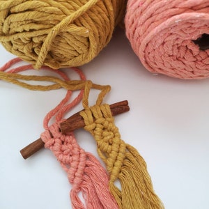 Puede incluir: Dos borlas de macram&eacute;, una rosa y otra amarilla, est&aacute;n atadas a un palo de canela. Las borlas est&aacute;n hechas con cord&oacute;n de algod&oacute;n grueso y tienen flecos largos y fluidos. Dos ovillos de hilo, uno rosa y otro amarillo, est&aacute;n en el fondo.