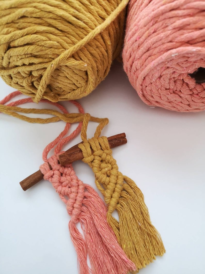 Puede incluir: Dos borlas de macram&eacute;, una rosa y otra amarilla, est&aacute;n atadas a un palo de canela. Las borlas est&aacute;n hechas con cord&oacute;n de algod&oacute;n grueso y tienen flecos largos y fluidos. Dos ovillos de hilo, uno rosa y otro amarillo, est&aacute;n en el fondo.