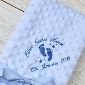 May include: A light blue baby blanket with a textured, dotted surface. The blanket features embroidered text and footprints in a darker shade of blue. A light blue satin ribbon and bow are attached to the bottom of the blanket. The text reads "Elliot James Marsh 13th January 2018."