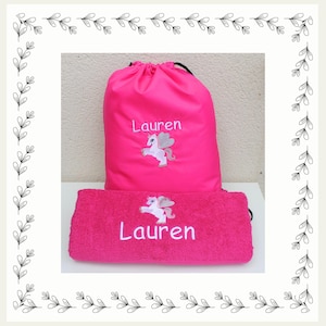 May include: A bright pink drawstring backpack and a matching pink rectangular towel. Both items feature an embroidered white unicorn with silver wings and the name Lauren stitched in white thread. The backpack has black drawstrings.